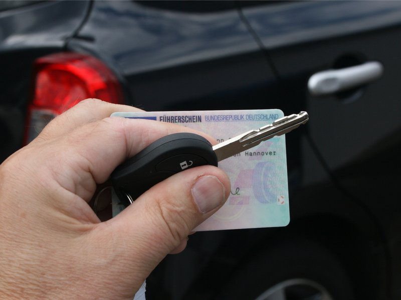 Eine Hand hält Autoschlüssel und Führerschein, den Hintergrund füllt ein schwarzer PKW aus. 